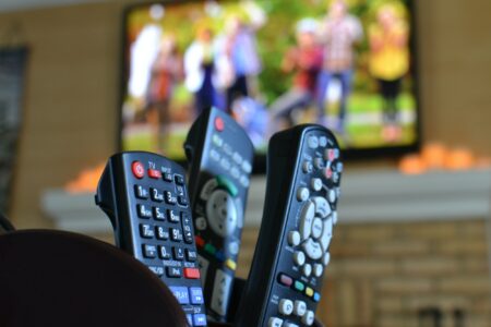 Tv Remotes With A Television With People Playing Football. Watching Movies, Sports Or Tv Shows